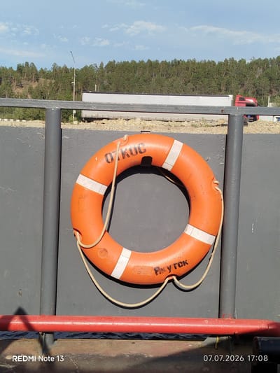 An orange life preserver with white stripes is hanging on a gray metal wall. The life preserver has black lettering on it which reads "ОТКРОЙ" on one side and "ЯКУТСЯ" on the other. A beige rope is attached to the life preserver and hangs down its side. In the background, a line of green trees stretches across a hill under a bright blue sky with scattered white clouds. A highway with trucks is visible in the distance. The bottom of the image shows a red metal railing. The bottom left corner of the image has the text "REDMI Note 13" and the bottom right corner has the date and time "07.07.2026 17:08". This scene appears to be located in Nizhny Bestyakh, Russia, as indicated by the text on the life preserver.