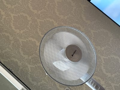The image shows a close-up view of a standing fan, with its metallic grid covering the fan blades. The fan is positioned in front of a wall covered with a patterned wallpaper. A portion of what appears to be a screen or monitor is visible in the upper right corner of the image, displaying a blue sky. The fan has a circular central hub with the logo "primera" visible on it. The base of the fan, with buttons, is also partially visible. The scene is indoors, suggesting a room in a building. The wallpaper has a damask-like pattern in muted tones of beige and cream. There are no people or animals visible in the image, and no specific actions are being depicted other than the static presence of the fan. The lighting suggests an indoor environment, possibly with natural light contributing to the illumination. There is no visual information in the image that can be used to determine its location in the city of Slonim, Belarus.