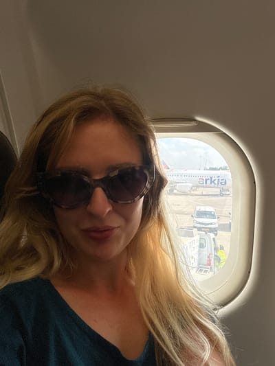 A woman with blonde hair and sunglasses is seated inside an airplane, looking at the camera. Outside the window, an Arkia airplane is visible on the tarmac, along with airport vehicles and personnel. The scene suggests the location is at Ben Gurion Airport in Yehud-Monosson, Israel, a major international airport serving the Tel Aviv metropolitan area.
