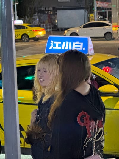 The image depicts a nighttime street scene in Longta, China. In the foreground, two young women stand near a yellow taxi. The woman on the left has blonde hair with bangs and is looking to her right with her mouth slightly open, as if speaking. The woman on the right has brown hair and is facing away from the camera, with her arm around the blonde woman's shoulders. Both women are wearing dark clothing.
Behind the women, a yellow taxi is parked, identified by a blue illuminated sign on its roof displaying Chinese characters and the word "TAXI". Another yellow taxi is visible further back on the left side of the image. A white car is parked to the right of the yellow taxi. The background shows a dimly lit street with building facades and streetlights. The overall impression is of a casual encounter on a city street at night.