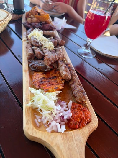 The image depicts an outdoor dining scene in Dobra Voda, Montenegro, featuring a large wooden platter of grilled meats and sides on a dark wooden plank table.

On the prominent light-colored wooden platter, a substantial assortment of grilled items is arranged. At the top, a pile of roasted or fried potato wedges is visible. Below this, various pieces of grilled meat include numerous small, elongated cylindrical items (likely ćevapi), several thicker grilled sausages, two longer pieces of grilled meat on skewers, and a large, flat piece of grilled meat or cheese with reddish seasoning and char marks. A mound of shredded, light-colored cheese or kajmak is placed over some of the ćevapi in the upper-middle section of the platter. At the bottom of the platter, from left to right, are distinct piles of shredded white cabbage, finely diced red onions, and a vibrant red relish or sauce (likely ajvar).

To the right of the wooden platter, a stemmed glass, characteristic of a wine or cocktail glass, holds a bright red/pink beverage with a dark straw protruding.

The table beneath the platter and drink is composed of dark brown wooden planks. In the upper left background, a portion of a woven basket is visible, along with a small, clear glass bottle and a dark-colored cup or container. Further in the background, out of focus, human hands are visible, one holding a fork over a white plate, indicating at least two individuals are present, one possibly wearing a red or striped garment. The overall lighting suggests a bright, possibly sunny, outdoor environment.