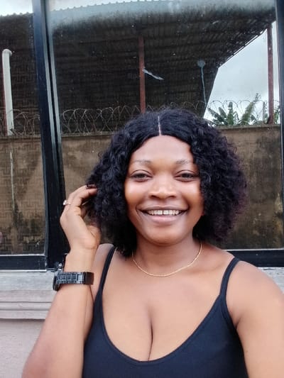 An adult female subject is depicted in a smiling close-up portrait, situated outdoors in Igbede, Nigeria. She has dark skin, black curly hair parted in the center, and is wearing a black sleeveless top, a thin gold-colored chain necklace, a dark-colored wristwatch on her left wrist, and a light-colored ring on her left ring finger. Her left hand is raised, with fingers touching the hair above her left ear. She smiles directly at the camera, revealing both upper and lower teeth. The background features a large glass panel or window that reflects an outdoor scene, including a corrugated metal roof structure supported by dark beams, a concrete wall topped with multiple strands of barbed wire, green foliage, and an overcast sky. A white vertical pipe is partially visible in the reflection on the left. A light-colored concrete ledge runs below the glass panel.