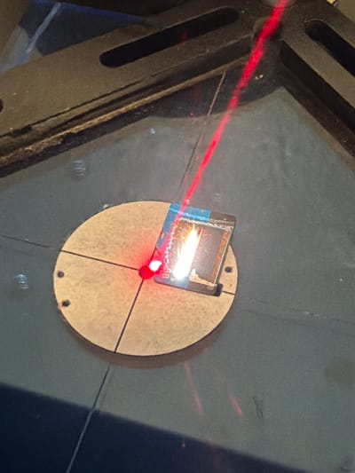 In Grapevine, Texas, a close-up, high-angle shot captures a detailed view of a scientific experiment. A small, dark square object, possibly a semiconductor chip, is positioned on a light-colored, circular platform marked with intersecting lines. A bright red laser beam is directed at the chip, creating a small, intense point of light and a visible beam path extending upwards and to the left. The chip appears to be encased in a blue and metallic material, with some signs of wear or damage. The circular platform is set against a dark, textured surface, suggesting a laboratory or workshop environment. The overall impression is one of precision and focused activity within a controlled setting.