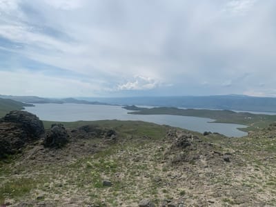 The image depicts a vast, panoramic view of a large body of water, likely Lake Baikal, with rolling, green hills and rocky terrain in the foreground. The sky is cloudy with patches of blue visible. In the distance, mountains form a hazy, blue horizon. The foreground is characterized by rough, stony ground sparsely covered with dry grass and small, white wildflowers. Several large, dark rock formations are prominent on the left side of the frame. The water stretches out, with peninsulas and islands dotting its surface. The scene conveys a sense of natural grandeur and remoteness. The location is identified as near Khuzhir, Russia. There are no people or animals visible in the image.