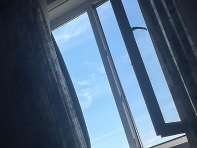 The image shows a view through a window in Gari, Russia. The window is partially open, revealing a bright blue sky with wispy clouds. Dark curtains, patterned with what appears to be a floral design, frame the window on both sides, with the left curtain drawn back to allow more light in. The window frame is a light gray color, and a handle for opening or closing the window is visible on the right side. The scene appears to be indoors, looking out onto an outdoor environment.