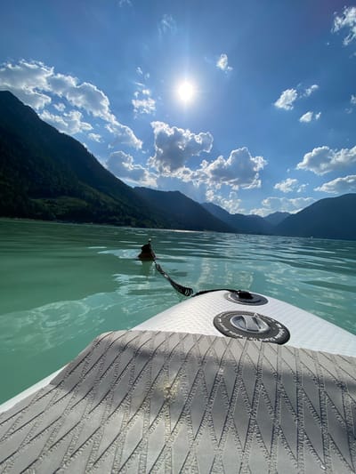 The image shows a first-person perspective of someone paddleboarding on a calm, clear body of water. The water is a turquoise green color, and gentle ripples are visible. To the left of the frame, a dark, tree-covered mountain range rises dramatically, with its slopes illuminated by the sun. The sky above is a vibrant blue, dotted with fluffy white clouds. The sun is bright and high in the sky, suggesting it is midday.

In the foreground, the edge of a white paddleboard is visible, with a textured grey grip pad. A black coiled leash is attached to a metal D-ring on the paddleboard and disappears into the water. The leash appears to be securing the paddleboard to the rider's ankle. There are no people clearly visible in the image, but the perspective suggests someone is actively participating in the activity. The scene evokes a sense of tranquility and enjoyment of the outdoors. The presence of mountains and clear water suggests a natural, picturesque location, consistent with the provided location context of Lärchenwiese, Austria. Text on the paddleboard is partially visible and reads "Inflatable Stand Up Paddle Board".