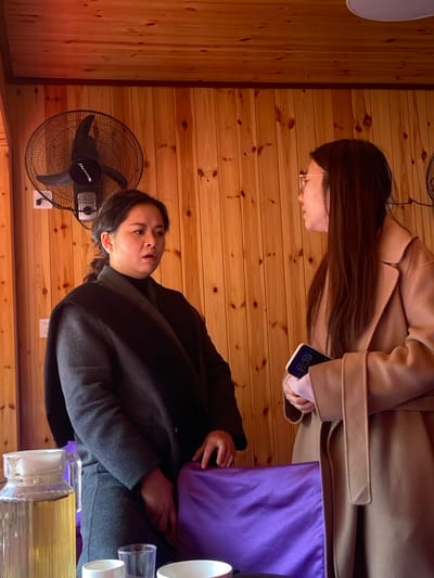 Two women are seated in a room with wood-paneled walls. One woman, with long brown hair, wears a tan coat and looks at her phone. The other woman, with shorter dark hair, wears a dark jacket. A fan stands against the wall behind her. A purple cloth and various containers are on a table near the women.