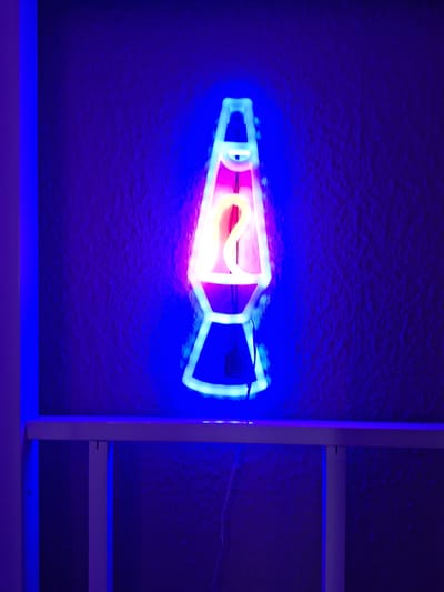 The image depicts a neon sign shaped like a lava lamp, illuminated against a textured dark blue wall. The neon tubing outlines the iconic shape of a lava lamp, with a bulbous top, a cylindrical middle, and a wider base. The colors of the neon are primarily bright pink and yellow for the "lava" within the lamp, and white and blue for the outer casing. The sign is positioned centrally in the frame, casting a vibrant glow on the surrounding area. A white shelf or ledge is visible in the foreground, below the neon sign. A thin black wire, likely part of the sign's power supply, dangles down from the base of the lava lamp neon and hangs near the shelf. The overall ambiance is dim and atmospheric, dominated by the intense colors of the neon light. The image does not contain any information that would allow for its location to be identified in the city of Wildeshausen, Germany.