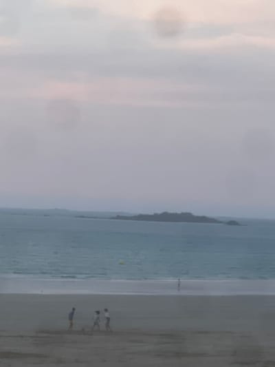 The image depicts a hazy, overcast day at a beach, likely in Pléneuf-Val-André, France, given the context. In the foreground, three indistinct figures are present on the sandy beach, appearing to be engaged in some activity, possibly playing. Further out, on the water's edge, a single, very small figure is visible near the surf. The sea is a muted blue-green with gentle waves. In the distance, a dark, rocky island or peninsula juts out from the horizon. The sky is a pale, diffused gray with hints of pink or lavender, contributing to the overall soft and indistinct quality of the scene. A yellow buoy is visible in the water between the shore and the distant island.