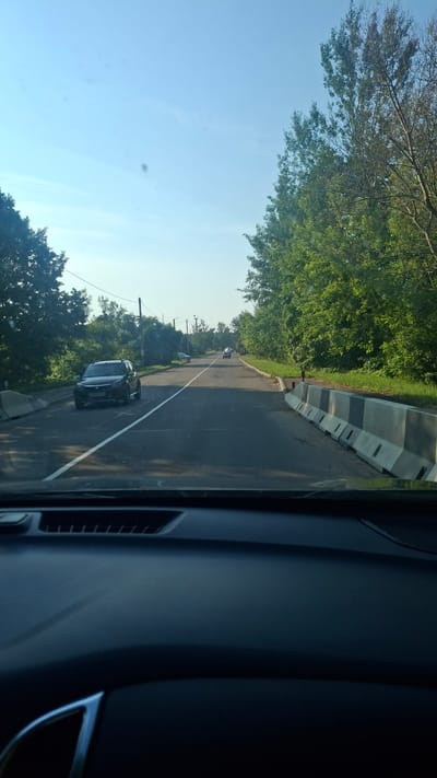 The image, taken from the perspective of a vehicle's dashboard, shows a two-lane road bordered by trees and concrete barriers. A dark-colored SUV is visible on the left side of the road, with its front wheels slightly turned. In the distance, other vehicles are present on the road. The sky is clear and blue. The scene appears to be on the outskirts of Stowbtsy, Belarus, indicated by the presence of the road, trees, and barriers which are common in rural or suburban settings in the region. The lighting suggests it is daytime.