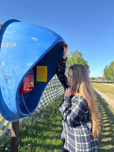 In Ust'-Silayka, Russia, a young female stands outdoors, using a public payphone on a bright, sunny day. She is positioned next to a large, vivid blue, shell-shaped payphone enclosure, which is supported by a rusty metal pole. Yellow digits "213111" are painted on the left exterior surface of the blue structure. The female has long, straight light brown hair and is dressed in a plaid flannel shirt featuring dark blue, green, and white checks. Her right hand grips the upper edge of the blue enclosure, while her left hand holds a red payphone receiver to her left ear. Her gaze is directed forward and slightly downward, indicating concentration.

Within the blue enclosure, a red payphone unit is mounted, displaying a silver-toned numeric keypad. To the right of the phone, a yellow rectangular note is affixed to the interior wall, containing dense Cyrillic text and what appear to be Braille-like tactile dots beneath some lines.

The surrounding environment depicts a rural landscape under a clear blue sky. A light-colored wooden lattice fence runs immediately behind the payphone on the left. A dirt road extends into the distance, curving to the right. The ground is covered in green grass, dotted with both dandelion flowers and seed heads. In the background, several deciduous trees with light green leaves are visible, along with distant wooden houses and utility poles. Long shadows are cast across the grass, consistent with late afternoon sunlight.