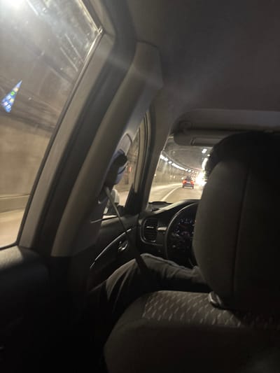 The image shows the interior of a car driving through a tunnel. The perspective is from the back seat, looking towards the front. A person's head and shoulders are visible in the foreground, seated in the driver's position. The steering wheel, dashboard, and part of the driver's legs are also visible. The tunnel is illuminated by overhead lights, and another car is visible ahead in the same lane. The exterior of the tunnel walls is visible through the side windows. There is no indication of the city of Kepsha, Russia, in the image.