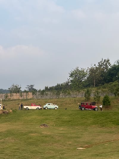 This image captures a serene outdoor scene in Babakan Madang, Indonesia, featuring a collection of vintage vehicles on a grassy hillside. In the foreground, the manicured green lawn slopes gently, dotted with a few scattered rocks. Beyond this, a short bamboo fence marks the edge of the property, separating it from a dense cluster of trees that form the backdrop.
The main subjects are three classic cars arranged on the slightly elevated ground behind the fence. On the left, a two-tone red and white pickup truck is parked. Next to it, a pale mint green sedan is positioned. To the right, a deep red vintage car with a distinct grille and fenders stands alone. Several individuals are visible around the vehicles, seemingly interacting with them or posing for photos. On the left, near the pickup truck, a group of at least four people are gathered. Near the red car on the right, two people are present, one of whom appears to be holding a camera.
The sky above is a pale, overcast blue with faint white clouds, suggesting a mild or hazy day. The overall impression is one of a carefully curated outdoor space, possibly a park or a venue designed for events or leisure, showcasing vintage automobiles against a backdrop of natural Indonesian landscape.