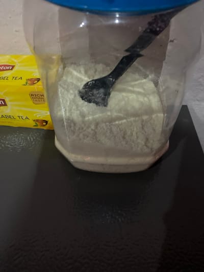 An indoor scene in the city of Mape, Nigeria, depicts a clear plastic container with an octagonal base, filled approximately one-third to one-half with a white powdered substance. A black plastic scoop rests partially submerged within the powder. To the left of the container, two yellow boxes of Lipton Yellow Label Tea are stacked horizontally, displaying brand logos, "Yellow Label Tea," and "RICH NATURAL TASTE." All items are situated on a dark, reflective surface, against a plain, light-colored background wall.