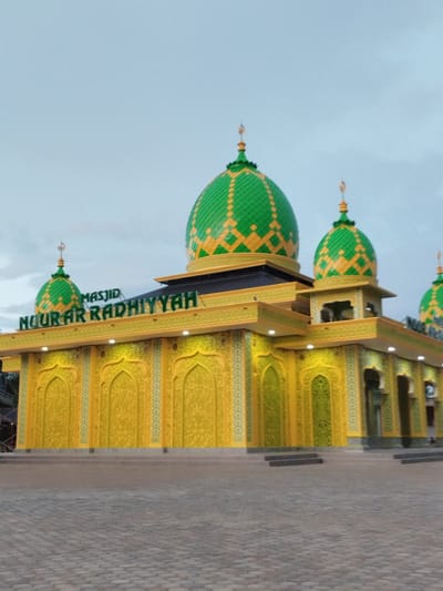 This full-shot image captures the vibrant Masjid Nuur Ar Radhiyyah in Aluekeujruen, Indonesia, under an overcast sky. The mosque features a prominent central green dome adorned with yellow diamond-shaped patterns, flanked by several smaller domes of similar design, each topped with a golden finial. The building's facade is predominantly golden yellow, intricately decorated with recessed arched panels featuring elaborate floral or geometric patterns in a lighter green-yellow tone. Green lettering on the front of the mosque clearly spells out "MASJID NUUR AR RADHIYYAH." Small white lights are visible along the building's eaves. In the foreground, a spacious courtyard paved with gray textured stones leads up to the mosque's entrance via multiple steps. No discernible individuals are present in the immediate vicinity.