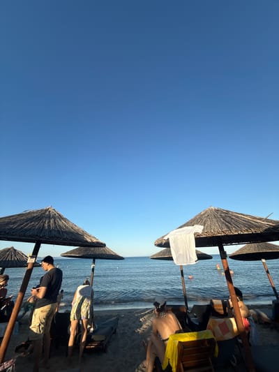 The image depicts a beach scene with people relaxing under thatched umbrellas. The setting is a sandy beach adjacent to the calm blue sea, bathed in the warm light of what appears to be late afternoon or early morning.

The subjects are primarily people, some of whom are seated or lying down on loungers, while others are wading in the shallow water. Two people are prominently visible near the foreground, one wearing a black t-shirt and light-colored shorts, looking down at a phone, and another person in a bikini, partially obscured. Further back, other individuals can be seen enjoying the water. Objects present include wooden posts supporting the umbrellas, loungers, and a white cloth draped over one of the umbrella structures. The umbrellas themselves are made of natural straw material, typical of beachside establishments.

The activity taking place is leisure and relaxation on a beach. People are sunbathing, swimming, and generally enjoying a day by the sea. The clear blue sky suggests good weather conditions, likely warm and sunny. The presence of multiple umbrellas and loungers indicates a developed beach area, possibly a resort or a well-equipped public beach.

Notable details include the clear, bright sky and the tranquil water. The orientation of the image is such that the sky occupies a large portion of the left side, while the beach and sea are on the right. The lighting suggests a time of day when shadows are not particularly long, hinting at a mid-day or late-day sun.

There is a small sign visible on one of the wooden posts, but the text is too small to be legible. The overall mood of the image is one of peace and relaxation, characteristic of a beach vacation. The location context of Nea Potidea, Greece, places this scene within a Mediterranean coastal environment.