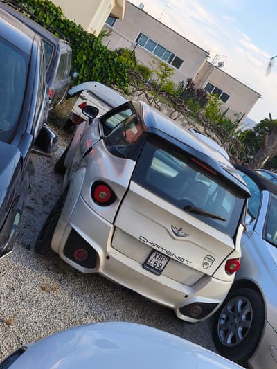 A white Chatenet microcar is parked on a gravel surface in front of a building in Perugia, Italy. The car's rear is visible, showing its distinctive taillights, a rear spoiler, and a license plate that reads "X8P L69". A black SUV is parked to the left of the microcar, and a silver car is parked to its right. The building behind the cars appears to be residential, with windows and a balcony. Green ivy covers part of the building and a chain-link fence. The sky is blue with scattered clouds. The overall impression is of a casual parking lot scene.