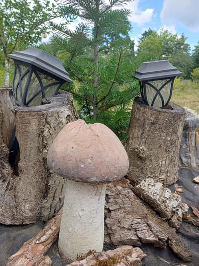 The image depicts an outdoor scene with a decorative mushroom sculpture in the foreground. The sculpture has a light brown, rounded cap and a textured, light-colored stem. Behind the mushroom, two tree stumps are visible. A black solar-powered lantern with a decorative glass enclosure sits atop each stump. The left stump is wider and has a hollowed-out section. The background features a young evergreen tree with green needles, and further back, mature deciduous trees and a blue sky with scattered clouds. The ground around the mushroom and stumps is covered with dark fabric and scattered pieces of bark. The scene is located in Sulymivka, Ukraine.
