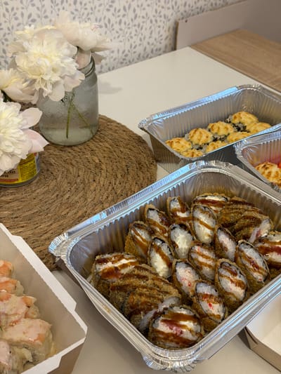 This media file shows a close-up, overhead view of several foil containers filled with sushi rolls. The primary subjects are the sushi rolls themselves, with distinct types visible. One container, on the left, holds rolls that appear to be baked or topped with a golden, toasted sauce and possibly sesame seeds. The other, larger container on the right, features deep-fried sushi rolls drizzled with a dark sauce, likely teriyaki or a similar glaze. The filling of these rolls is visible, showing creamy white ingredients, possibly cream cheese, and some red or pink elements, suggesting seafood.
In the foreground and to the left, there are several white peony flowers, some in a clear glass jar filled with water, suggesting a decorative element in a domestic or restaurant setting. A textured, woven placemat is also visible under the jar and part of the sushi container. A small, cylindrical can with visible text is partially obscured by the placemat and flowers.
The overall scene suggests a meal, likely a takeaway or delivery, presented for consumption. The lighting appears to be natural or from an indoor source, casting soft shadows. The focus is on the food, highlighting its presentation and variety. No people are visible in the frame.
The visible text on the can appears to be in Cyrillic, with words like "ВЯЛЕНЫХ" and "ПОРОВ" partially decipherable. Based on the context of sushi and the visible text, the location is likely Russia. The time of day and weather are not discernible from the image. The emotion conveyed is one of appetizing presentation and potential enjoyment of the meal.