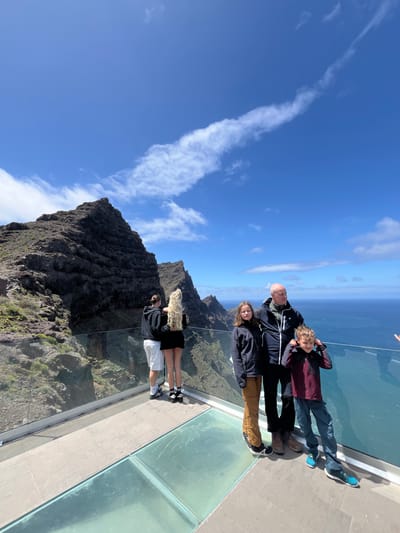 The image captures a family of five standing on a glass-bottomed viewing platform overlooking a mountainous landscape and the ocean. The location is in La Aldea de San Nicolás, Spain. The family consists of two adults, a teenager, and two younger children. They are positioned along the edge of the platform, seemingly enjoying the view. The backdrop features rugged mountain peaks with sparse vegetation, a clear blue sky with streaks of white clouds, and the vast expanse of the ocean. The platform is constructed with glass panels, providing a transparent view of the terrain below.