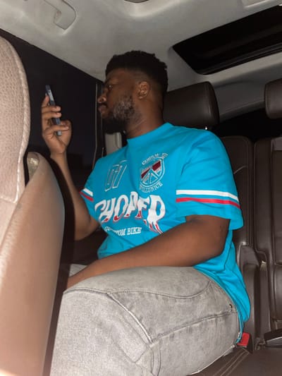 A man with dark skin, short dark hair, and a beard is lying down in the back seat of a vehicle. He is wearing a blue t-shirt with a white and red design and gray jeans. He is holding a mobile phone in his left hand. The interior of the car includes leather seats and light-colored trim. The location appears to be Jiru, Nigeria.