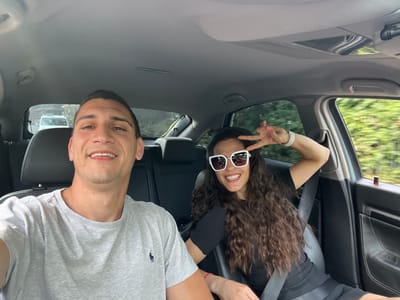 Two individuals are visible inside a car. A woman with curly brown hair and white sunglasses is making a peace sign. She is wearing a seatbelt and a black shirt. A man wearing a gray t-shirt is next to her. Both are smiling. The car is in motion, and trees are visible through the car windows. Another car is visible through the back window. The exact location in the city of Maidenhead, United Kingdom, is not identifiable.