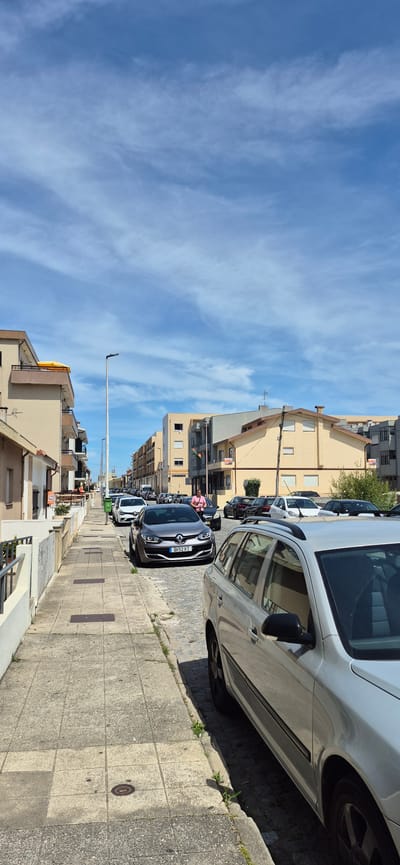The image shows a street in Vila do Conde, Portugal, with several cars parked along the side. The street is paved with cobblestones and has a sidewalk on the left side. The buildings on either side of the street are two to three stories high and appear to be residential. The sky is blue with some white clouds. There is one person walking on the sidewalk, and another person in a pink shirt standing by a car. The time of day appears to be daytime, with sunlight casting shadows.