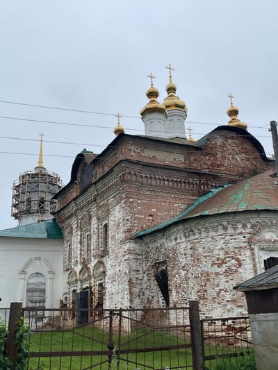 The image shows the exterior of an old, weathered, brick church in Kstinin, Russia. The church features multiple golden domes topped with crosses, a characteristic of Orthodox Christian architecture. The brickwork is a mix of red and white, with significant signs of age and decay, including peeling paint and exposed brick. Some sections of the church appear to be under repair, with scaffolding visible around a tall, slender dome. The sky is overcast, creating a muted lighting condition. In the foreground, a rusty metal fence runs across the scene, with green grass visible behind it. A small, nondescript structure with a slanted roof is partially visible on the right side of the image. The overall impression is one of historical significance and a sense of ongoing preservation efforts for the ancient religious site.