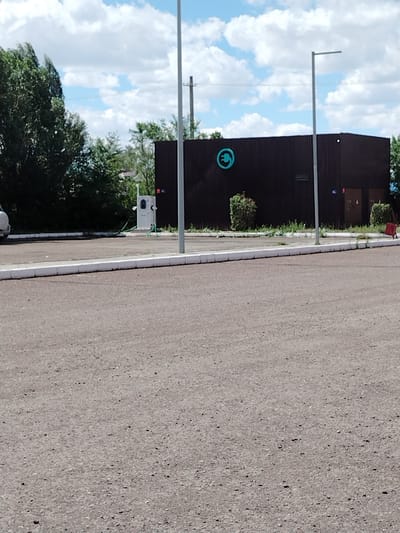 The image depicts an outdoor scene, likely in the city of Ekibastuz, Kazakhstan, focusing on an electric vehicle charging station. A dark-colored, corrugated building with a circular electric plug logo is visible in the background. To the left of the building, a white electric charging station is situated, with a green charging cable extending from it onto the asphalt. A portion of a white vehicle is parked to the far left. The foreground is dominated by a paved area, possibly a parking lot, with a white curb marking its edge. Trees and a bright blue sky with scattered clouds complete the environment.