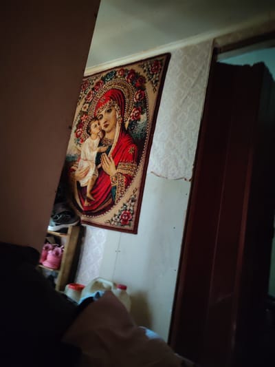The media file is a dimly lit, medium-angle shot of an indoor scene, likely a residential setting in the Voskresensky District of Russia. The primary subject is a framed tapestry hanging on a wall, depicting a religious icon of the Virgin Mary holding the infant Jesus. The tapestry is ornate, featuring rich red and floral patterns. To the left, a dark, indistinct object, possibly furniture or a partition, partially obscures the view.
In the background, a doorframe with a dark, possibly wooden door is visible. The wall adjacent to the tapestry appears to be wallpapered with a subtle pattern, but a section near the top is peeling away, revealing a lighter surface underneath.
In the lower left of the frame, shelves are visible with several items, including a pair of athletic shoes and what appear to be pink child-sized boots. Further to the right, on the floor or a low surface, are two large plastic containers with red caps, possibly for water or other liquids. A crumpled piece of fabric, possibly bedding or clothing, is also present in the foreground.
The lighting is low, suggesting it might be evening or a poorly lit room. There are no obvious external weather cues. The overall atmosphere appears somewhat cluttered and domestic. No visible text is present in the image. The scene does not depict any specific activity or event, but rather a static display of personal belongings and decor.