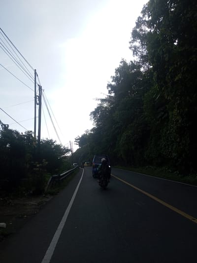 The photograph depicts a paved road cutting through a verdant landscape. The scene is possibly located in the outskirts of Phou Khoun, Laos, evidenced by the presence of dense foliage and a winding road common in mountainous regions. A motorcycle with two riders follows a blue van, while other vehicles are farther ahead. Power lines stretch across the sky on the left, supported by utility poles. The sun is bright and partly obscured by thin clouds. The right side of the road is lined with a dense forest, while a guardrail and some vegetation are present on the left. The asphalt has a single yellow line down the middle and a white line on the left edge. The image is taken from behind the motorcycle.