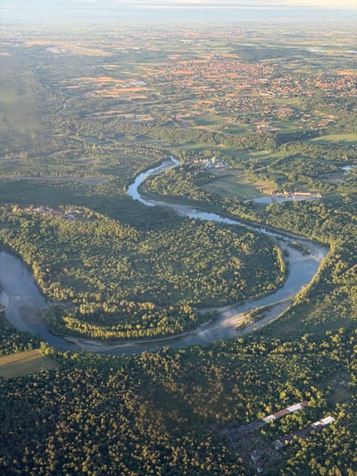 This aerial photograph presents a vast landscape combining natural features with human development, indicative of the region around Pombia, Italy. The primary subject is a wide, light blue river, likely the Ticino, which meanders significantly through the scene, flanked by extensive, dense green forests. Patches of lighter sand or gravel banks are visible along the river's edges.

To the left of the main image, and more distinctly in the first crop, a large residential area spreads across the landscape. Numerous houses with reddish-brown roofs are clustered together, forming a town or village. Adjacent to and interspersed with these settlements are various agricultural fields, appearing as rectangular patches of green and yellow, suggesting cultivated land.

Several man-made structures are visible within the forests. In the second and third crops, long, narrow buildings with light-colored roofs, possibly industrial facilities, warehouses, or large greenhouses, can be seen. One crop also shows a straight, dark line cutting through the landscape, which could be a road or a railway line.

The image suggests a time of day during the golden hour, either late afternoon or early morning, evidenced by the long shadows cast by trees and structures, and the warm, golden light illuminating parts of the terrain. The sky appears clear, though a light atmospheric haze is visible, particularly towards the horizon on the left. No specific human activity is discernible from this high-altitude perspective, nor is any visible text present. The scene conveys a sense of natural beauty and the subtle integration of human habitation within it.