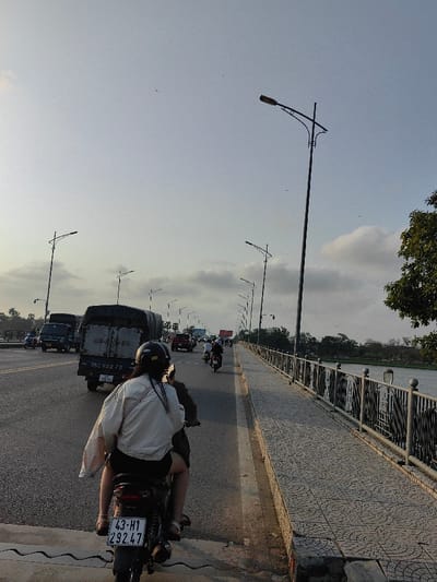 The image depicts a late afternoon or early evening scene on the Phú Xuân Bridge, spanning the Perfume River in or adjacent to Thuận Hóa Ward, Huế City, Thừa Thiên Huế Province, Vietnam.
In the foreground, a dark-colored Honda motorcycle, bearing license plate "43-H1 292.47," is observed from a rear perspective. It carries two occupants. The rear passenger, identified as female, wears a dark helmet, a light-colored, loose-fitting, long-sleeved shirt, dark shorts, and sandals. Her long dark hair is visible beneath the helmet, and she carries a light-colored bag over her left shoulder. The driver, largely obscured by the passenger, wears a dark helmet and dark clothing.
The motorcycle is positioned on a multi-lane asphalt roadway, with traffic flowing in the same direction. Immediately ahead of the motorcycle is a dark green utility truck, license plate "75C-122.70," featuring a canvas-covered cargo area. Further down the bridge, multiple other vehicles, including another similar utility truck, a red pickup truck, and several motorcycles, are visible in motion.
To the right of the roadway, a pedestrian and bicycle path, paved with gray interlocking tiles in a circular pattern, extends along the bridge. This path is bordered by a dark metal bridge railing, which incorporates decorative vertical panels featuring cutout designs. Beyond the railing, the calm surface of the Perfume River is visible, reflecting the pale sky. The distant riverbank is lined with green trees and foliage.
Overhead, a series of tall streetlights with distinct decorative arching fixtures line both sides of the bridge, extending into the distance. The sky is a pale blue-grey with scattered white cumulus clouds, consistent with twilight conditions. Numerous small dark specks, likely birds or insects, are visible against the sky. The streetlights are not illuminated. The overall environment indicates an active urban bridge crossing with moderate traffic.