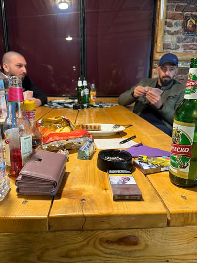 This image captures an indoor scene within an establishment in Mednikarovo, Bulgaria. Two adult males are visible seated at a light-colored wooden table. The individual on the left has a shaved head, a dark beard, and wears a dark long-sleeved garment, partially facing away from the camera. The individual on the right wears a blue baseball cap and a dark jacket, possesses a beard, and is actively holding and examining a hand of playing cards.

The wooden table is extensively covered with various items:
In the foreground right, a green glass bottle of Burgasko beer with a white and green label is prominently placed. To its left, a pack of Rothmans cigarettes lies next to a black ashtray containing several cigarette butts and ash. A yellow disposable lighter rests on a purple fabric napkin beside the ashtray, with a black pen positioned nearby. A 20-unit banknote is partially visible, folded, further to the left.
In the mid-ground, a purple zippered wallet rests on the table, adjacent to an opened bag of potato chips with yellow and red packaging. A white oval plate holds a grilled sausage and a metal fork.
In the foreground left, multiple bottles are present: a clear glass bottle with a pink label and white text (possibly a soda or mixed drink), another clear bottle, and a red-capped bottle, likely containing a condiment.
In the background on the table, two green bottles (likely beer) and an orange bottle are visible.

The background environment features a large, dark-red paneled wall, possibly a window or partition, showing reflections of overhead lights and indistinct figures. To the far right, a section of an exposed brick wall is visible, with a small framed religious icon mounted on it. The overall setting suggests a casual social gathering, possibly a bar or pub, where individuals are engaging in a card game while consuming beverages and snacks.