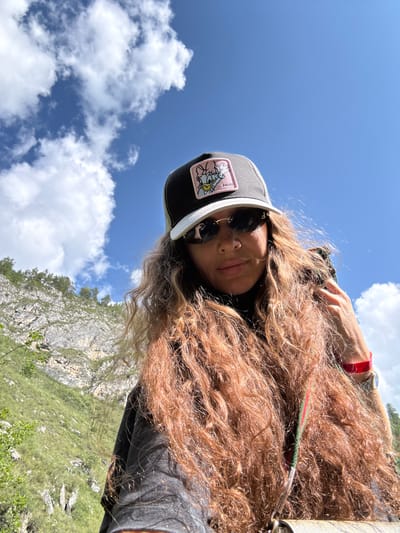 A woman with long, curly auburn hair is taking a selfie outdoors under a blue sky with scattered white clouds. She is wearing a black baseball cap with a patch featuring Daisy Duck, dark sunglasses, and a black shirt. A red wristband is visible on her right wrist, and she appears to be wearing a watch. Her left hand is holding a strap over her shoulder. The background shows a steep, rocky hillside covered in green vegetation, suggesting a natural, possibly mountainous, environment. The specific location in Chemal, Russia is not determinable from the image.