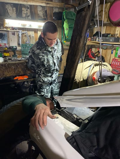 In the city of Kauchuk Stantsiya, Russia, within a dimly lit garage, two men are engaged in work on a vehicle. The garage's interior is rustic, with wooden plank walls and a visible beam overhead. One man, wearing a camouflage jacket, stands and appears to be observing the work. The other man, wearing a dark green hoodie and a baseball cap, is positioned in front of the vehicle, leaning over its engine compartment. His hands are actively engaged, suggesting he is performing repairs or maintenance. The vehicle's hood is open, revealing the engine components. Various tools and equipment are scattered around the garage, including a toolbox, a drill, and orange work gloves on a workbench. The overall scene depicts a common, practical activity within a utilitarian setting.