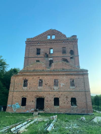 The image shows an abandoned, multi-story brick building in Yuryatino, Russia. The building has a damaged roof and several missing windows. There is graffiti on the exterior walls, and the ground in front of the building is overgrown with grass and debris. The sky is clear blue.