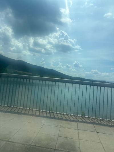 The image shows a view from a bridge over the Pravets Reservoir in Pravets, Bulgaria. A metal railing runs horizontally across the foreground, casting shadows on the paved walkway. Beyond the railing, the reservoir's calm blue water stretches towards a lush green hillside covered in trees. The sky above is a mix of bright blue and dramatic, dark clouds, with sunlight breaking through in places. The overall scene depicts a scenic natural landscape under a dynamic sky.