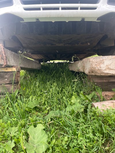 The image shows the underside of a white vehicle parked on concrete blocks in a grassy area. The vehicle's undercarriage is dirty and appears to be a car or SUV. The concrete blocks are rough and weathered, and they are stacked on wooden planks. The ground is covered in dense green grass and weeds, with some larger leaves visible. The scene appears to be outdoors, possibly in a rural or suburban setting. There are no people visible in the image. The location is identified as Nizhnii Bugrysh, Russia.