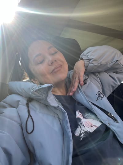 A female individual is observed seated inside a vehicle, assumed to be in the city of Sel’khoztekhnika, Russia. The scene is characterized by intense sunlight entering from the upper left, generating visible lens flares. The individual's eyes are closed, head slightly tilted back, and lips parted. She wears a light grey puffer jacket, partially unzipped, revealing a dark grey t-shirt underneath which features a graphic print depicting a pale, classical-style figure. The jacket displays a black "The North Face" logo on the right chest area. Her right hand, bearing a thin ring on the ring finger, is positioned on the left side of her neck/upper chest. Dark hair is pulled back, exposing multiple small hoop earrings on the left ear. The interior of the vehicle, including a seat headrest and a portion of the roof lining, is visible in the background.