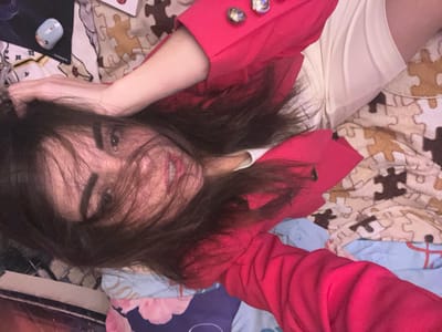 The image is a close-up, slightly tilted, indoor selfie of a young person lying down. The person has dark, slightly messy hair that partially obscures their face, and they are wearing a bright red jacket over a white top. The background consists of a bed with a duvet cover featuring a puzzle piece pattern in muted colors like beige, brown, and white, along with a blue sheet underneath. To the left of the frame, a small light blue computer mouse with a floral design is visible on a dark surface, which appears to be a mousepad with the word "SONNEN" printed on it. There is also a partially visible white object with a red circular design and some text, likely a book or packaging, in the upper left corner. The lighting is soft and appears to be coming from above and to the left. There are no elements in the image that provide a specific location within Krasnoe-na-Volge, Russia; the scene is generic and could be anywhere.