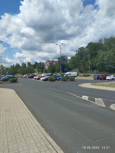 A daytime, eye-level shot captures a street scene in Karlovy Vary, Czechia. The foreground features a paved sidewalk leading to a grey asphalt road. Beyond the road, a parking lot is filled with various cars, predominantly sedans and some vans. Trees and buildings line the background, with some trees showing lush green foliage. The sky is a mix of blue and grey clouds, indicating partly cloudy weather. The image also includes traffic signs, streetlights, and a white van parked in the lot. A person in blue shorts is visible walking in the distance. The timestamp "18.06.2026 14:21" is present in the bottom right corner of the image.