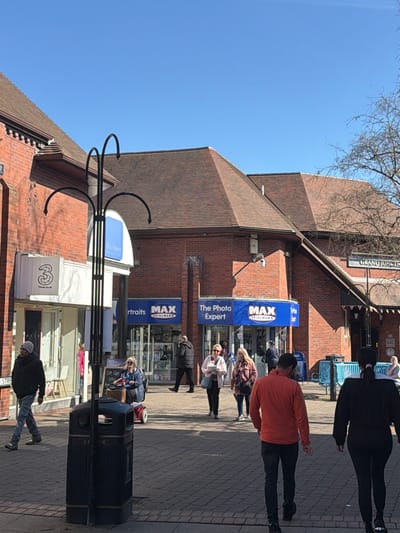 The image depicts a bustling pedestrianized street scene on Market Street in Crewe, United Kingdom, near the Grand Junction entrance to the Market Shopping Centre, under a clear blue sky.
The environment is characterized by predominantly red brick buildings with dark brown tiled roofs. On the left, a "3" (Three Mobile) store with a white fascia is visible. Further along the street, two "MAX SPIELMANN" photo processing shops are present, labeled "Portraits" and "The Photo Expert," both featuring blue fascias with white lettering. In the background on the right, a building with a "GRAND JUNCTION" sign suggests an entrance to a larger retail area. The pedestrian pathway is paved with dark grey block paving. A bare-branched tree is visible on the far right.
In the foreground, slightly left of center, a dark, ornate metal street furniture element with multiple curved arms stands in front of a dark grey litter bin.
Approximately 15-20 individuals are present. In the immediate foreground, facing away from the viewer, a male in an orange top and dark trousers walks on the right, alongside a female with dark hair in dark attire. In the mid-ground, walking towards the viewer on the left, is a male wearing a dark jacket and a grey beanie. A female in a blue jacket is seated on a red mobility scooter, equipped with a brown basket, facing right. Further back, a male in a dark coat walks past Max Spielmann. Other individuals, casually dressed, are walking along the street; a female with blonde hair carries a white item, and another with a patterned scarf walks nearby. Several people appear to be entering or exiting the shops. There are no clear direct interactions visible between individuals. Blue bollards or barriers are visible further down the street on the right. Security cameras are mounted on the building facades.