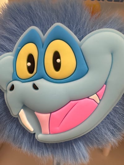 The image is a close-up shot of a blue furry creature's face. The creature has large, cartoonish yellow eyes with black pupils and white highlights. Its mouth is open, revealing a pink tongue and sharp white teeth. The creature's face is light blue with darker blue accents around its eyes. The background is blurred and appears to be a light brown surface. There is no visible context of Keila, Estonia in this image.