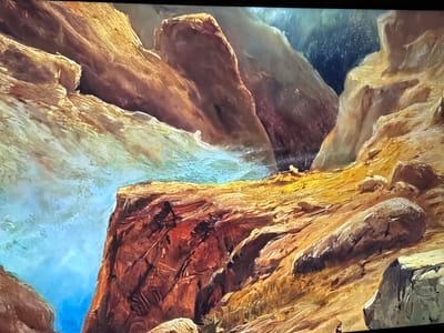 The image displays a rugged natural landscape, confirmed as being located in Reus, Spain. The scene is dominated by a deep gorge or canyon composed of layered rock formations with a distinct body of water flowing through it.

The rocks exhibit a varied palette of warm earth tones, including rich browns, reds, oranges, and golden yellows, particularly on illuminated surfaces. Darker, deep red-browns and shadowed areas reveal crevices and depths within the rock faces. Textures are predominantly rough and angular, with visible striations, cracks, and evidence of erosion, suggesting sedimentary rock. Some areas, particularly closer to the water's edge, appear smoother from water flow.

A vibrant body of water occupies the lower-left and central-left sections, characterized by striking turquoise and deep blue hues. White streaks and foamy patches indicate movement or currents within the water. The water's edge meets the rocky banks sharply.

In the upper-left, lighter, yellowish-brown rock formations slope towards the water. The upper-right background features darker, more obscured rock formations, some appearing almost black, possibly due to deep shadow or distant, dark vegetation. The mid-right foreground includes a large, elongated, light-colored geological feature resembling a fallen log or a distinctive rock formation, resting on a sandy-colored, sloping rock surface. The overall lighting is strong and directional, creating dramatic highlights and deep shadows that accentuate the three-dimensional complexity of the terrain. No human figures, animals, or man-made objects are visible.