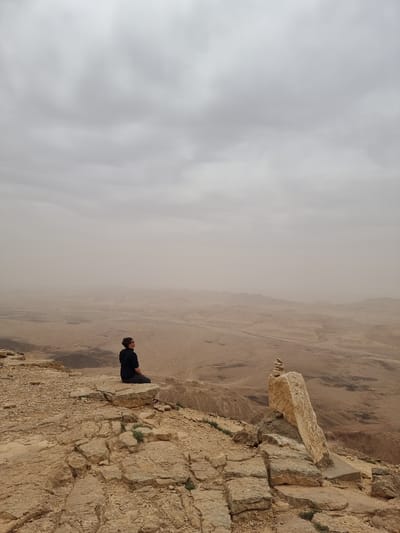 The image shows a man sitting on the edge of a cliff in Mitzpe Ramon, Israel. He is facing out towards a vast, arid landscape. The sky is overcast with heavy cloud cover. The cliff edge is rocky and irregular. In the background, the landscape stretches out in layers of brown and tan hues. A rock formation is visible to the left of the man.