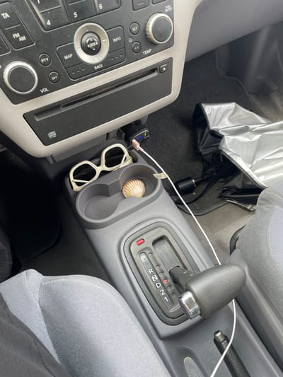 The image shows the interior of a car. The focus is on the center console area, which includes the gear shifter, cup holders, and part of the radio/infotainment system. In one of the cup holders, there is a small seashell. Next to it, a pair of white-framed sunglasses rests in another cup holder. A white charging cable is visible, connected to a car charger plugged into the dashboard.
To the left of the gear shifter, a portion of the car's control panel is visible, with buttons and knobs for various functions like radio tuning, volume, and temperature settings. Some text labels are visible, including "TA", "FM", "AM", "PUSH AUTO", "SET", "INFO", "MAP", "OFF", "VOL", and "disc CY460". The gear shifter itself displays the common automatic transmission settings: P, R, N, D, and numbered gears.
In the background, a dark-colored car floor mat is visible, along with a partially opened umbrella with a silver exterior and black interior, lying on the floor behind the center console. The seats of the car are upholstered in a gray fabric. The lighting suggests it is daytime, and the overall scene conveys a sense of everyday use and perhaps a touch of personal items brought into the vehicle, such as the seashell and sunglasses. The location context of La Línea de la Concepción, Spain, implies a warm climate, which might explain the presence of sunglasses.