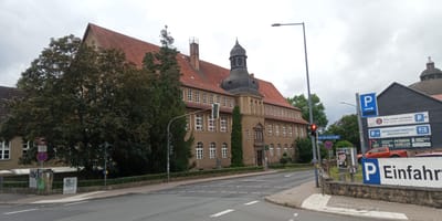 The image captures a street scene in Eisenach, Germany, featuring a large, historic building with a red tile roof and a dark, turreted section on the right. The building appears to be a school or administrative complex, with numerous windows. A traffic light is visible, currently showing red. Street signs indicate "Joh.-Seb.-Bach-Straße" (Johann Sebastian Bach Street) and a parking sign with a blue 'P'. There are trees lining the street and a large tree on the left side of the building. Several other signs are visible, including those for "Öffentlicher Parkplatz" (Public Parking) and various accommodations like "HÖSTEL & PENSION „ALTE BRAUEREI“" and "PENSION SCHÖLLER". An orange pickup truck is parked to the right. The sky is overcast.