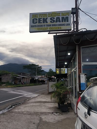 The image presents a quiet roadside scene in what appears to be a rural or semi-urban area, likely Sukamulia, Indonesia. The sky is heavily overcast, suggesting either late afternoon or early evening, with artificial lights already active on the building to the right.

A prominent rectangular sign hangs above this building, displaying text in Indonesian: "BEK TUWO KERIPIK DAPU CEK SAM." Below this, it reads, "HANYA DISINI !!! TIDAK BUKA CABANG LAIN" (Only here!!! No other branches opened) and "KHAS SAREE" (Saree specialty), indicating a business that specializes in crisps or chips. The building itself is a single-story structure with large windows, possibly a shop or restaurant, and has several pendant lights illuminating the area under its awning. A white car, partially visible, is parked in front of the building.

In the foreground, a damp asphalt road with a white dividing line runs alongside a concrete sidewalk. Across the road, a row of simple wooden shacks or houses can be seen. The background is dominated by a large mountain, its upper slopes and peak obscured by thick, low-hanging clouds, contributing to an atmospheric and somewhat muted scene. Trees are visible on the mountain and in the midground. Smaller, partially visible signs on the building's facade also mention "DUA PUTR," "MEU H MATA," and place names like "Medon" and "Saree." No people are visible, lending a tranquil and still quality to the image.