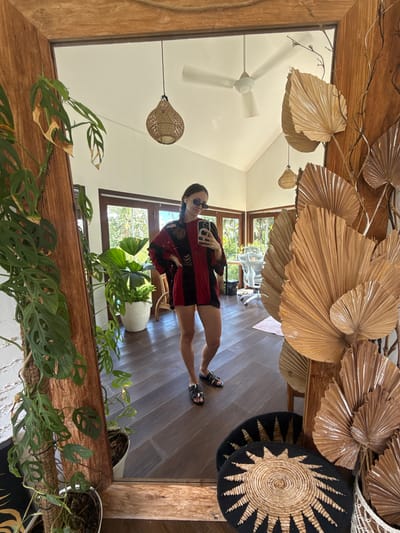 The image shows a reflection in a wooden-framed mirror. Reflected is a person wearing sunglasses, a red and black top, and short shorts. The person is holding a mobile phone, likely taking a selfie.
The reflection shows the interior of a room with a wooden floor. There is a ceiling fan. A variety of plants in pots are also reflected. Dried palm fronds are arranged above in an artful display. Woven wall hangings can also be seen.
Based on the decor and plant life visible, the image is likely taken in Mas, Indonesia, as indicated in the prompt.